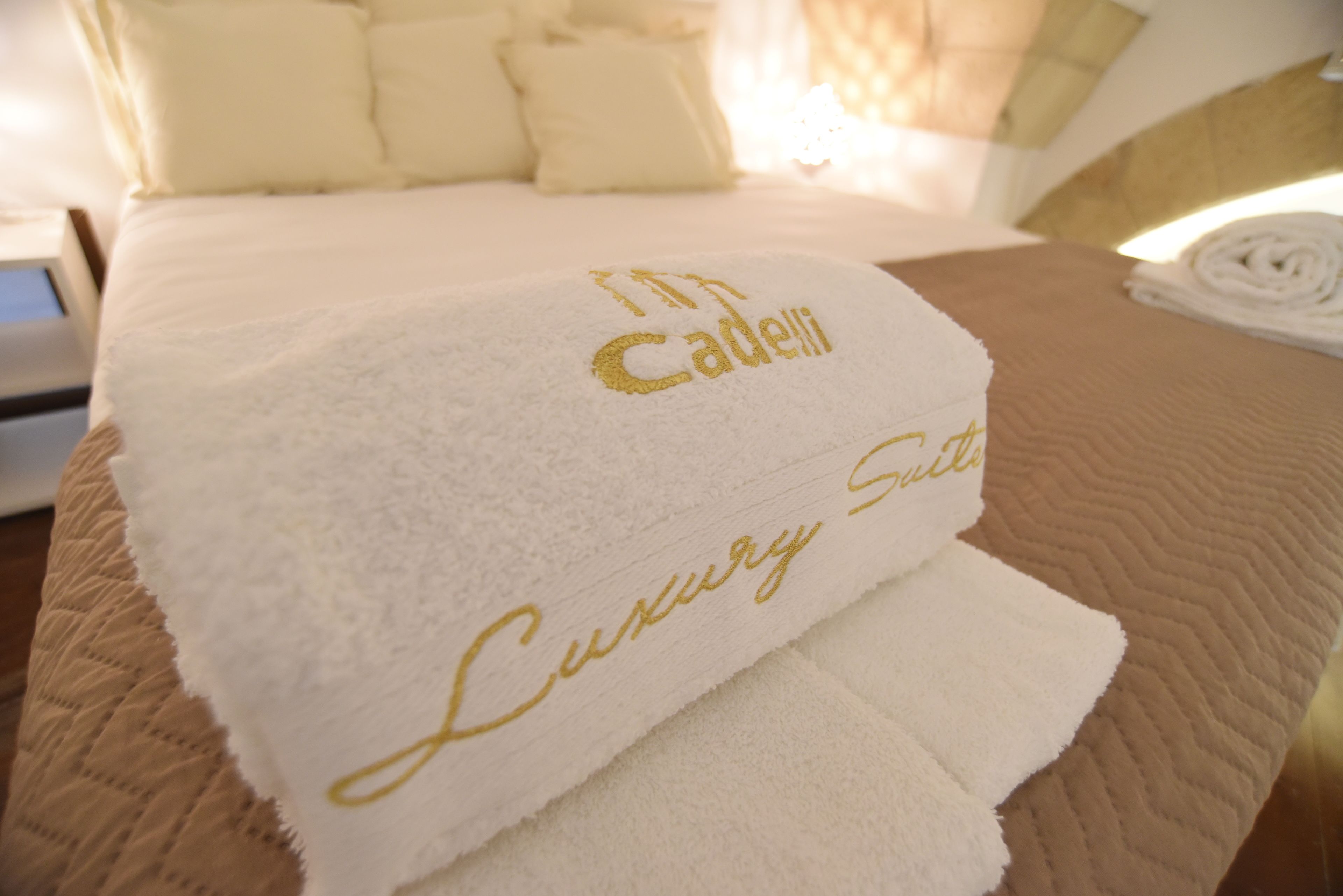 Een bed met een witte deken en een handdoek met de tekst 'Cadelli Luxury Suites' erin geborduurd.