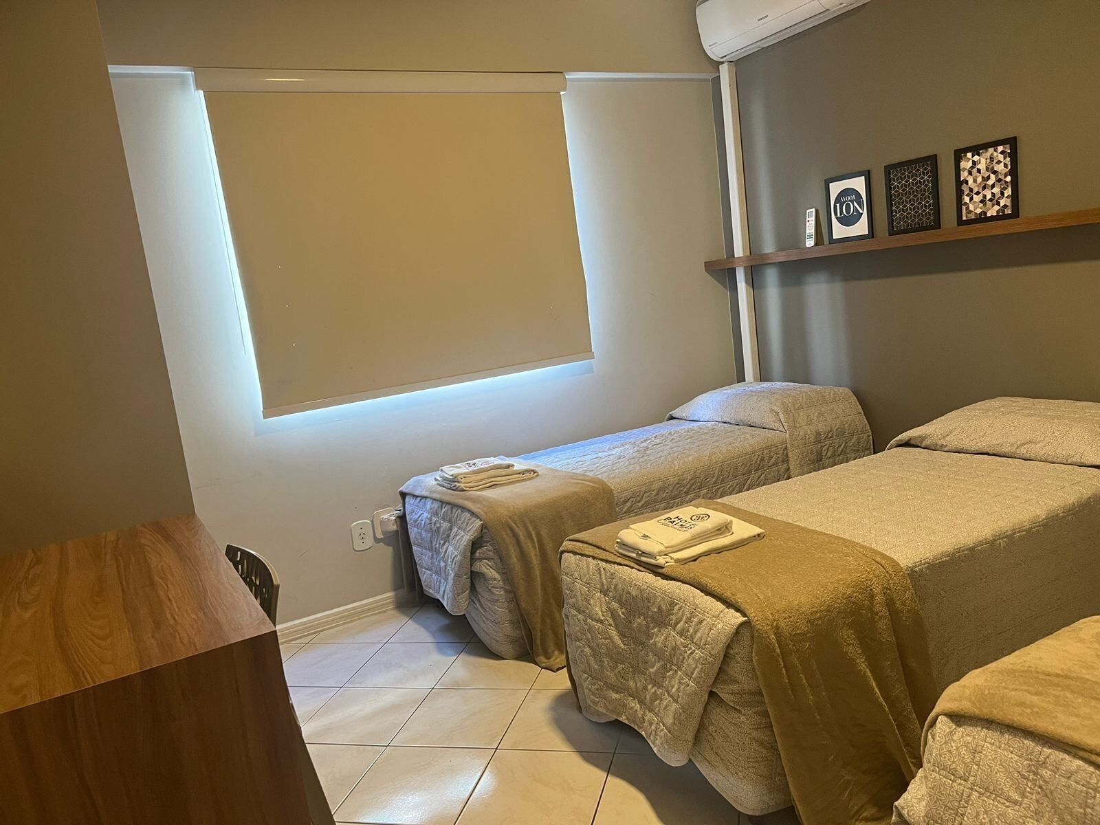 A hotel room with two beds, a window with blinds, a wall-mounted shelf with framed pictures, and a wooden desk.
