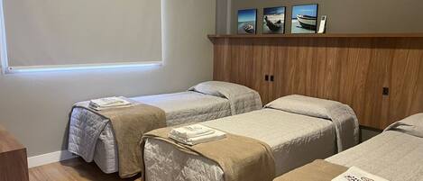 A hotel room with two beds, a wooden wardrobe, a window with blinds, and three framed pictures on the wall.