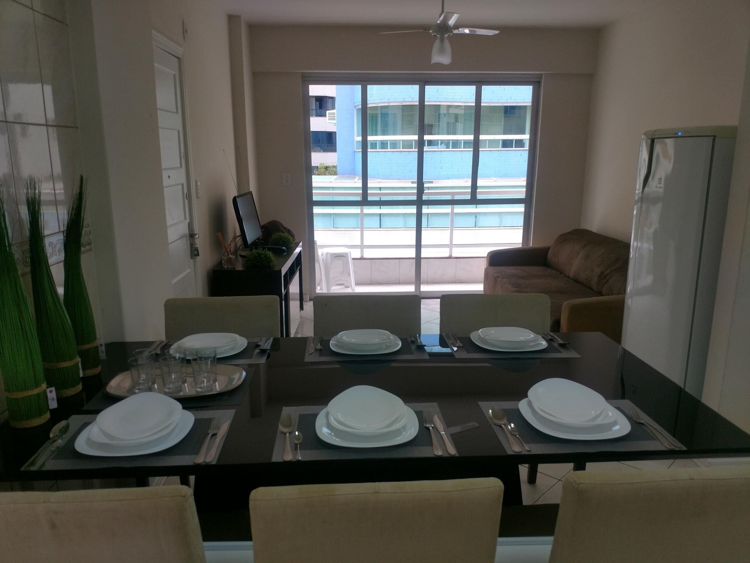 A dining area with a table set for four, a ceiling fan, and a view of a pool and buildings through large windows.