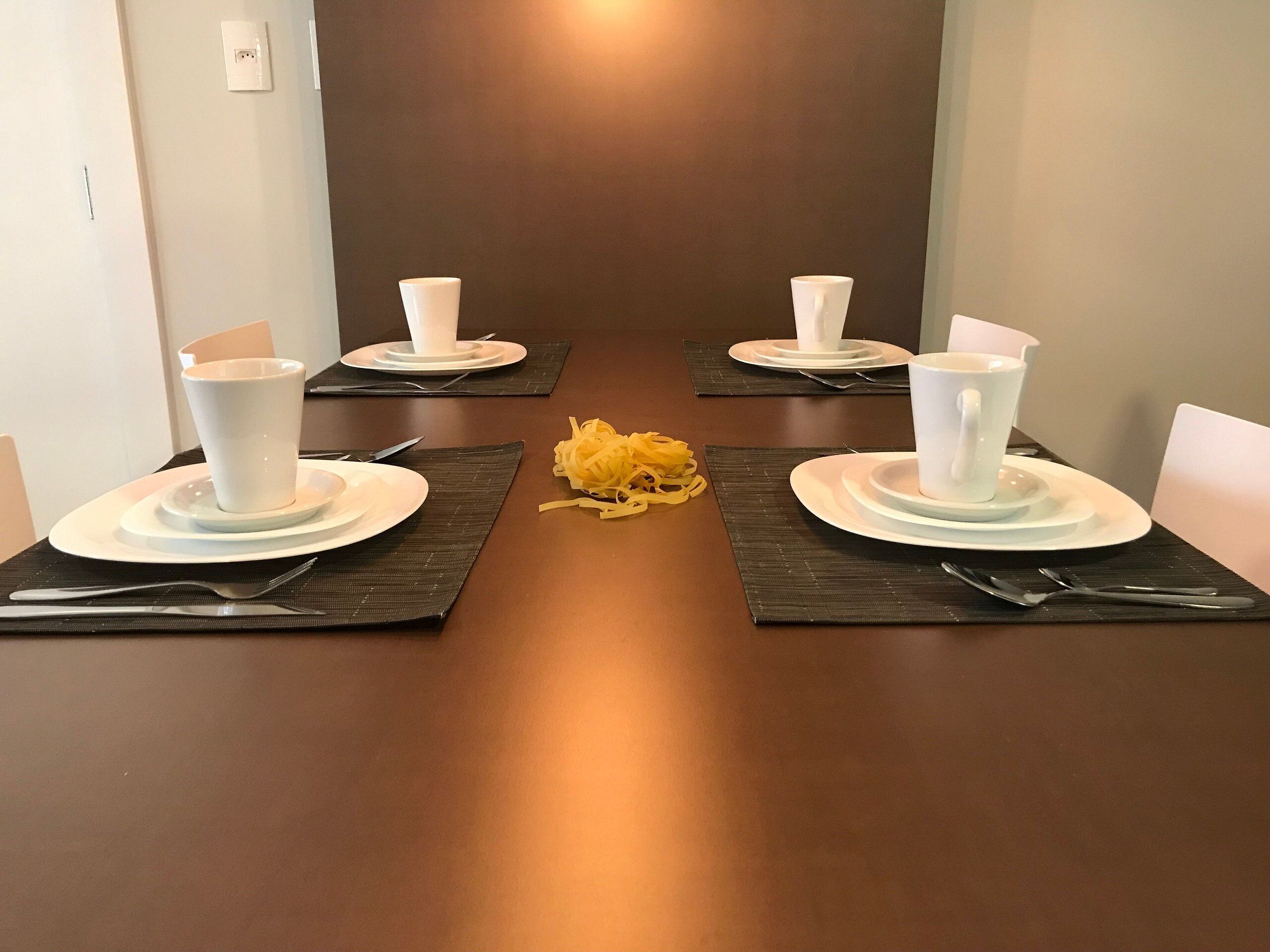 Een eettafel gedekt voor vier personen, met witte borden, bekers en bestek, en een decoratief geel accent in het midden.