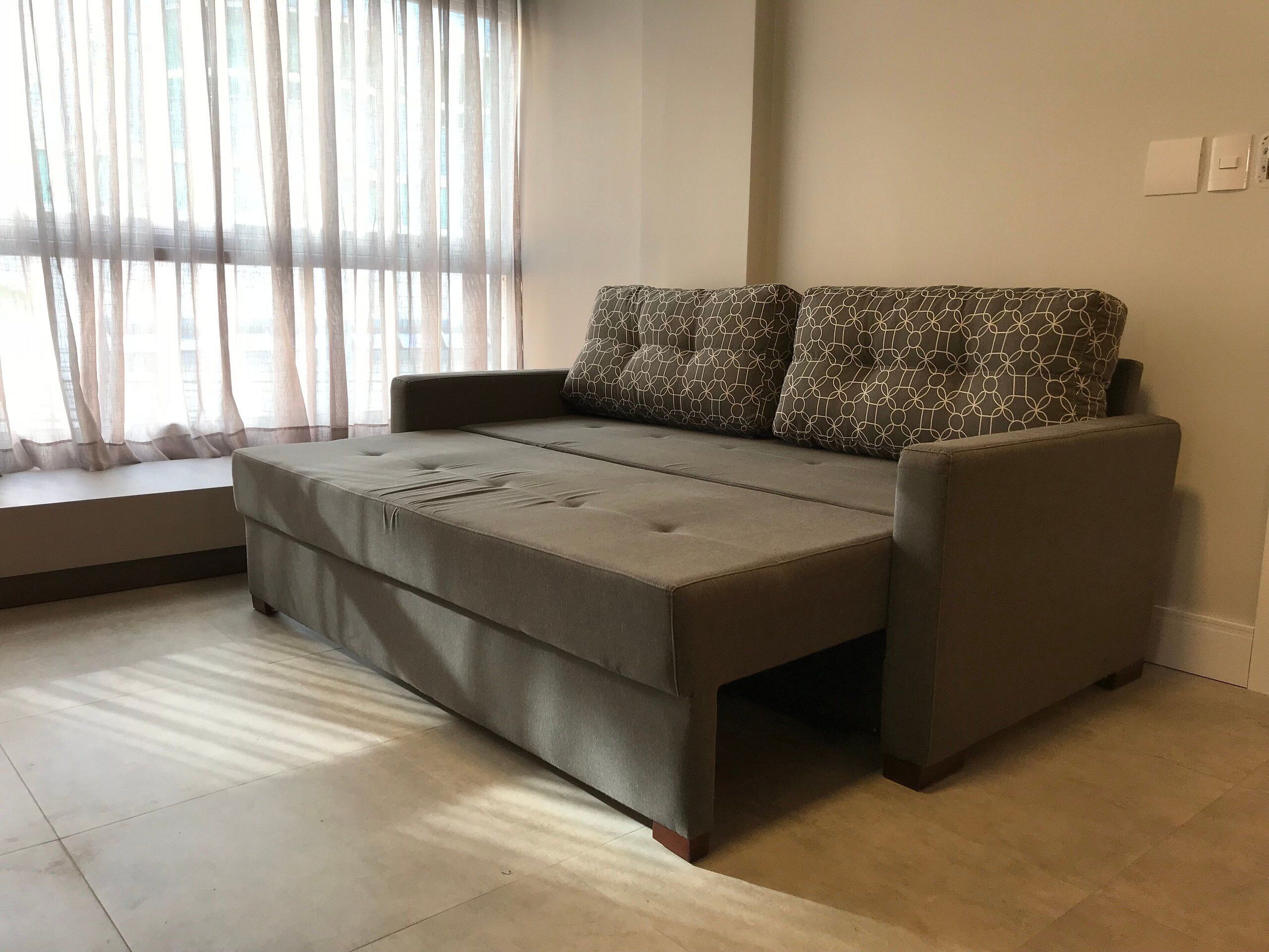 Una sala de estar moderna con un sofá y un reposapiés, un ventanal con cortinas ligeras y un piso de baldosas.