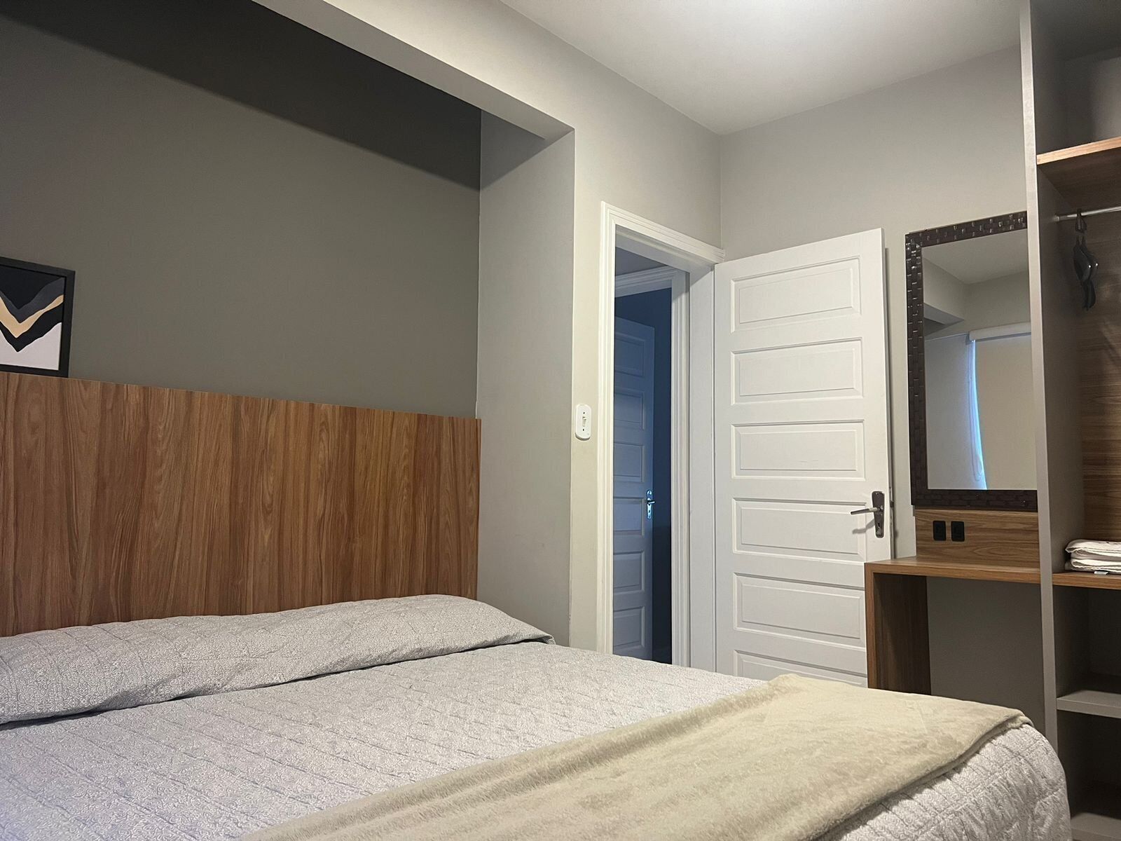A bedroom with a wooden headboard, a bed with a grey quilt, a white door, and a mirror on the wall.
