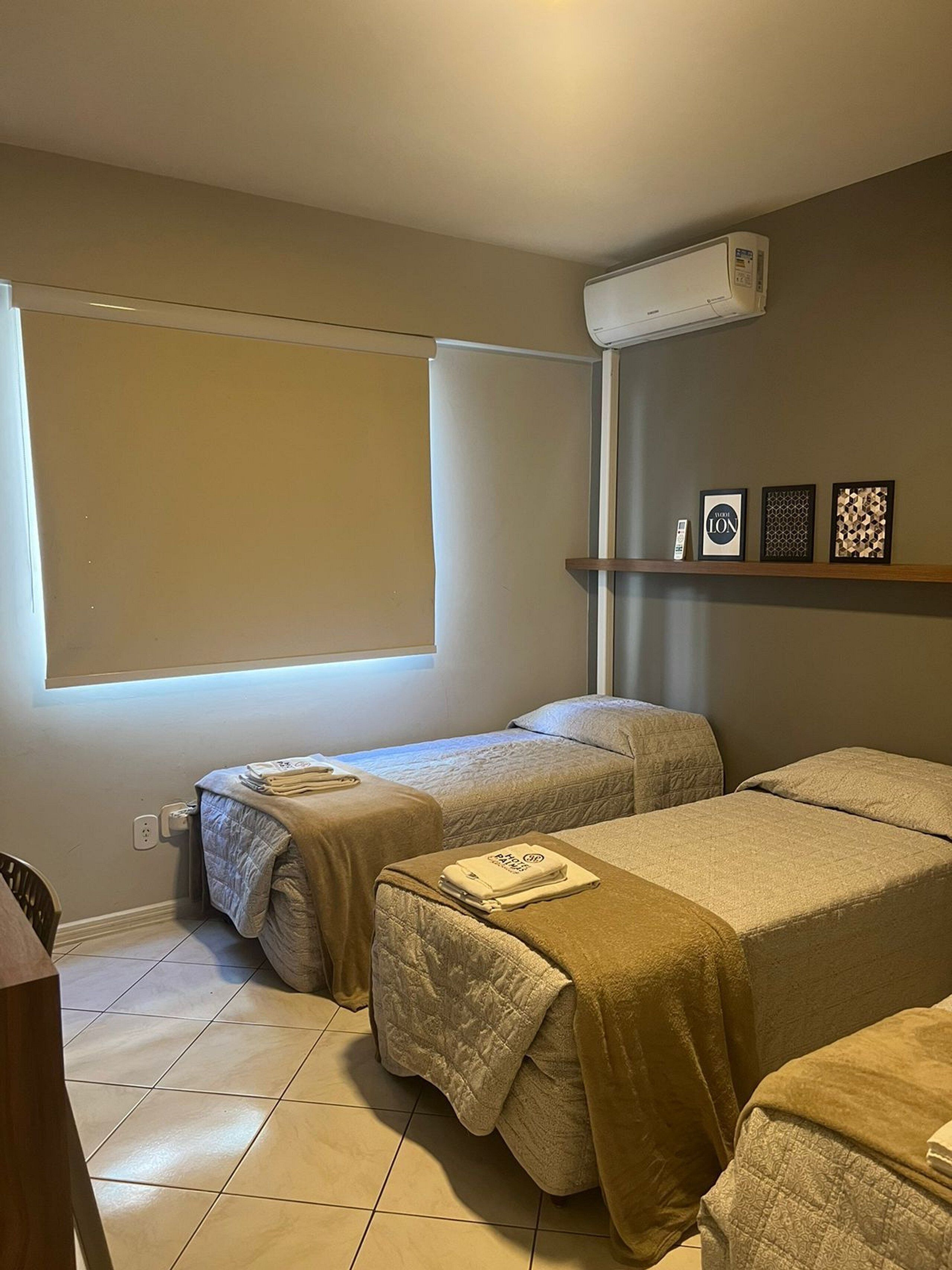 Habitació d'hotel amb dos llits, una finestra gran, aire condicionat i una estant amb quadres emmarcats.