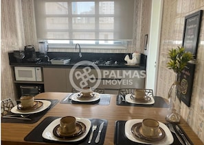 A dining area with a kitchen in the background, a window, and a sign for 'Palmas Imóveis'.