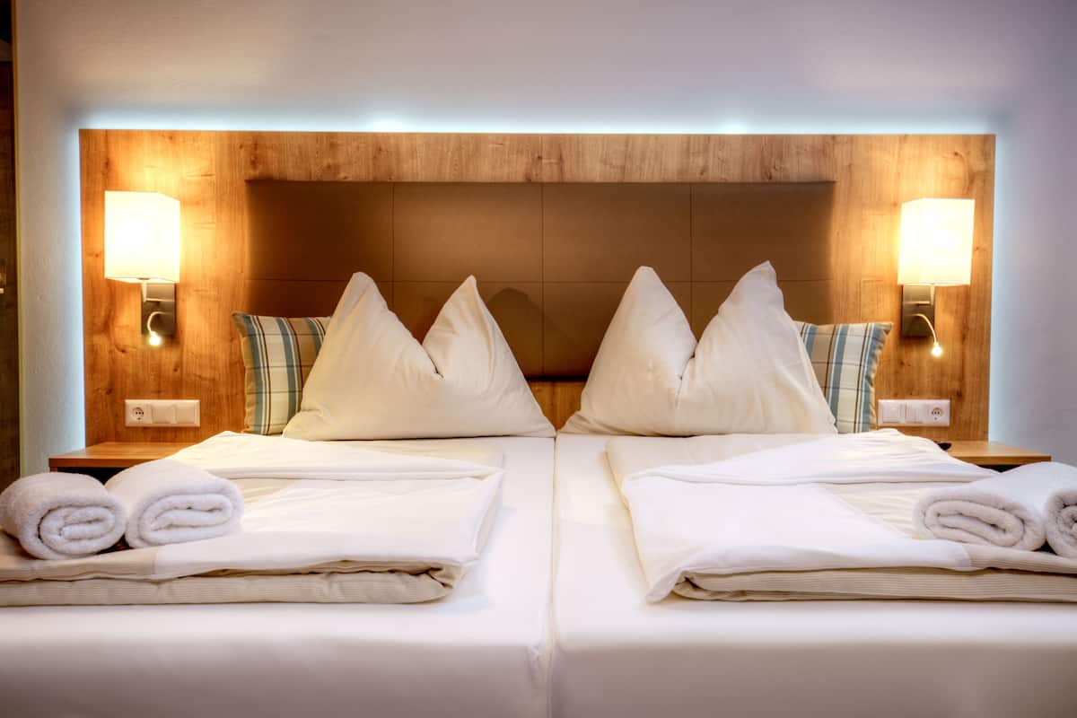 A hotel room with two beds, each with neatly folded white towels, wooden headboard, and wall-mounted lamps.