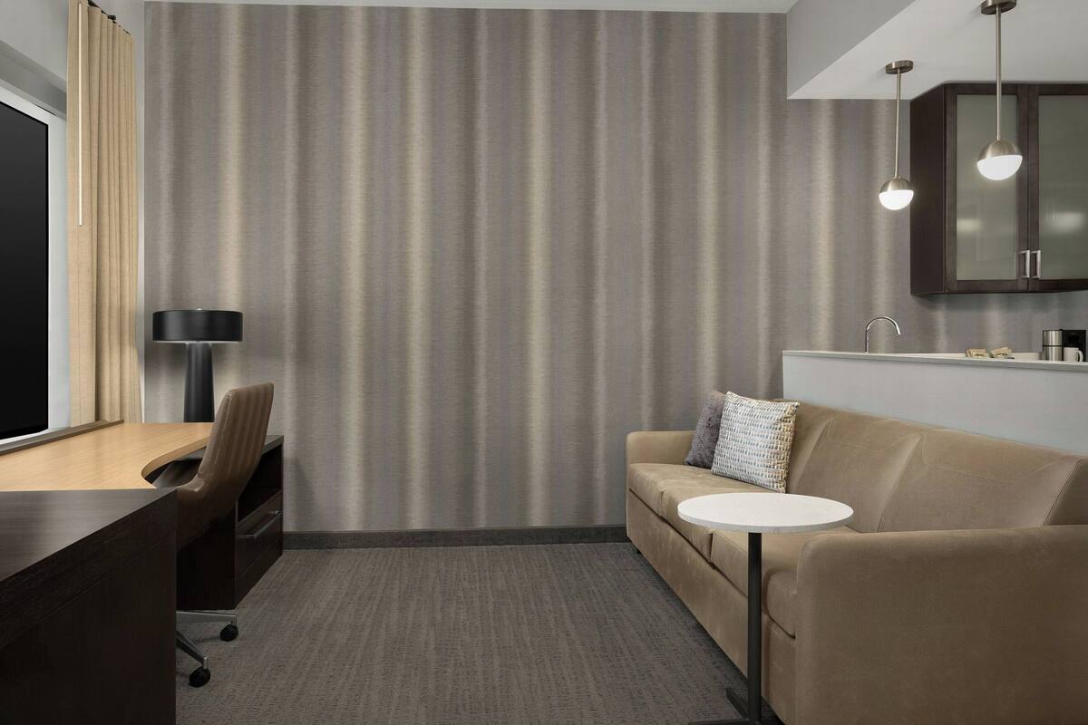 A modern hotel room with a desk, a sofa, a small round table, a lamp, and a cabinet.