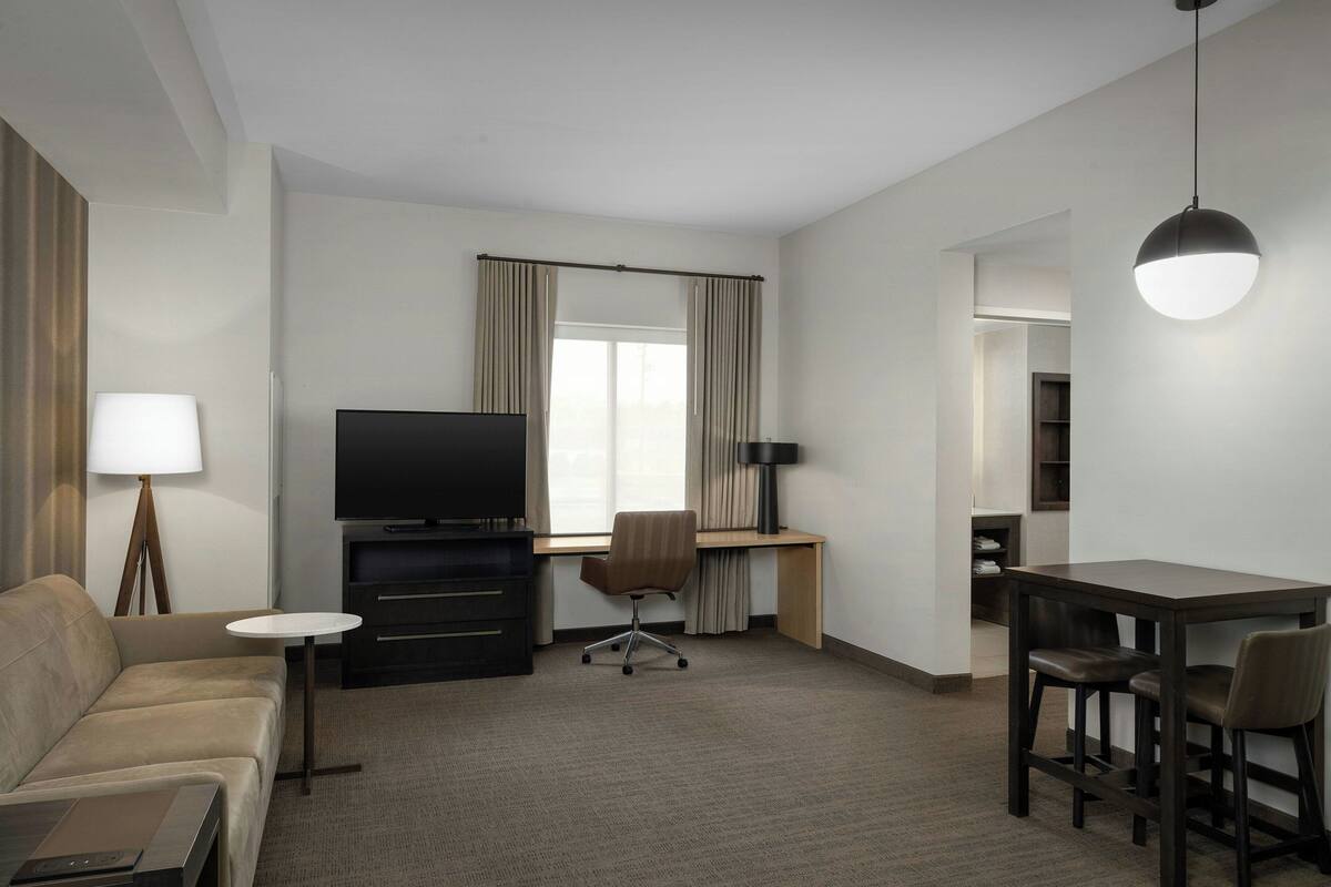 A hotel room with a sofa, a TV, a desk, a dining table with chairs, a lamp, and a window with curtains.