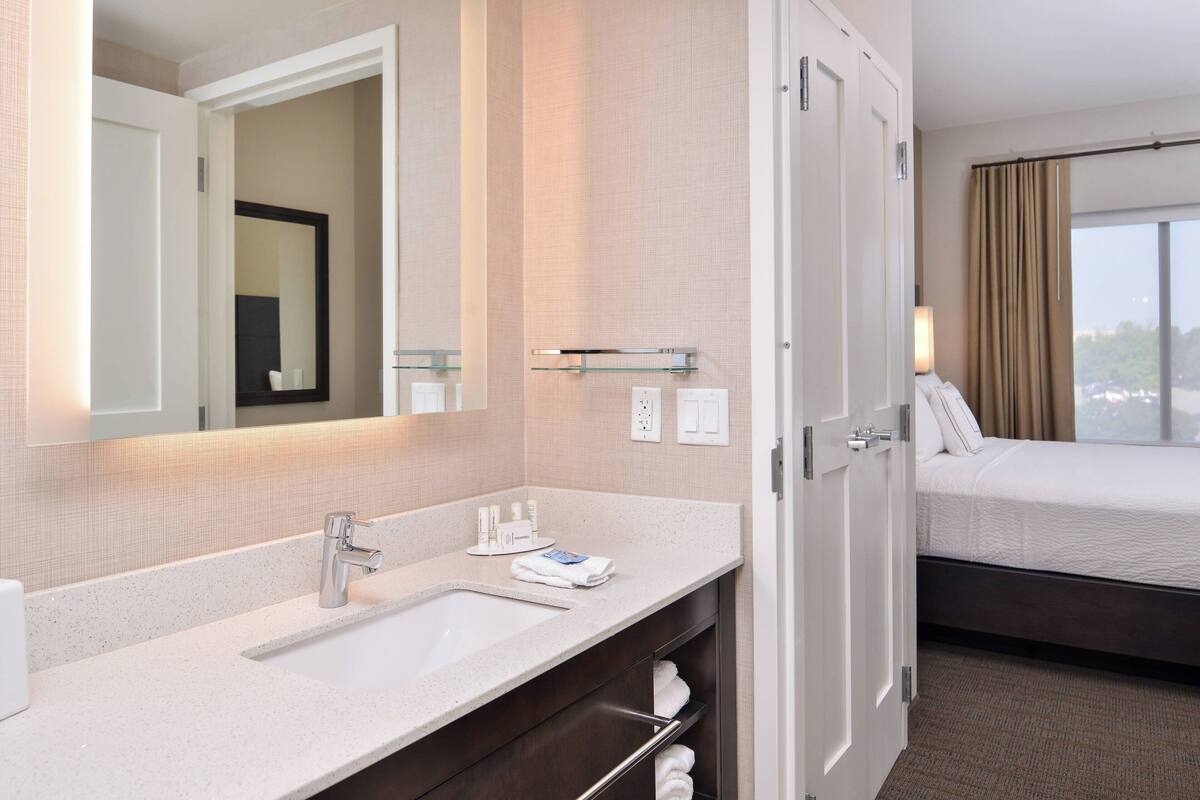 A hotel room with a bathroom featuring a sink, mirror, and towel rack. There is a bed with a window and curtains.