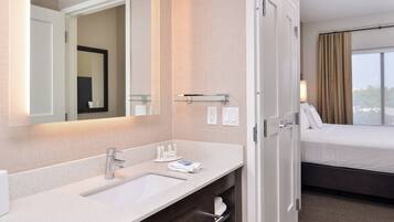Une chambre d’hôtel avec une salle de bain comprenant un lavabo, un miroir et un porte-serviettes. La chambre est équipée d’un lit, d’une fenêtre et de rideaux.