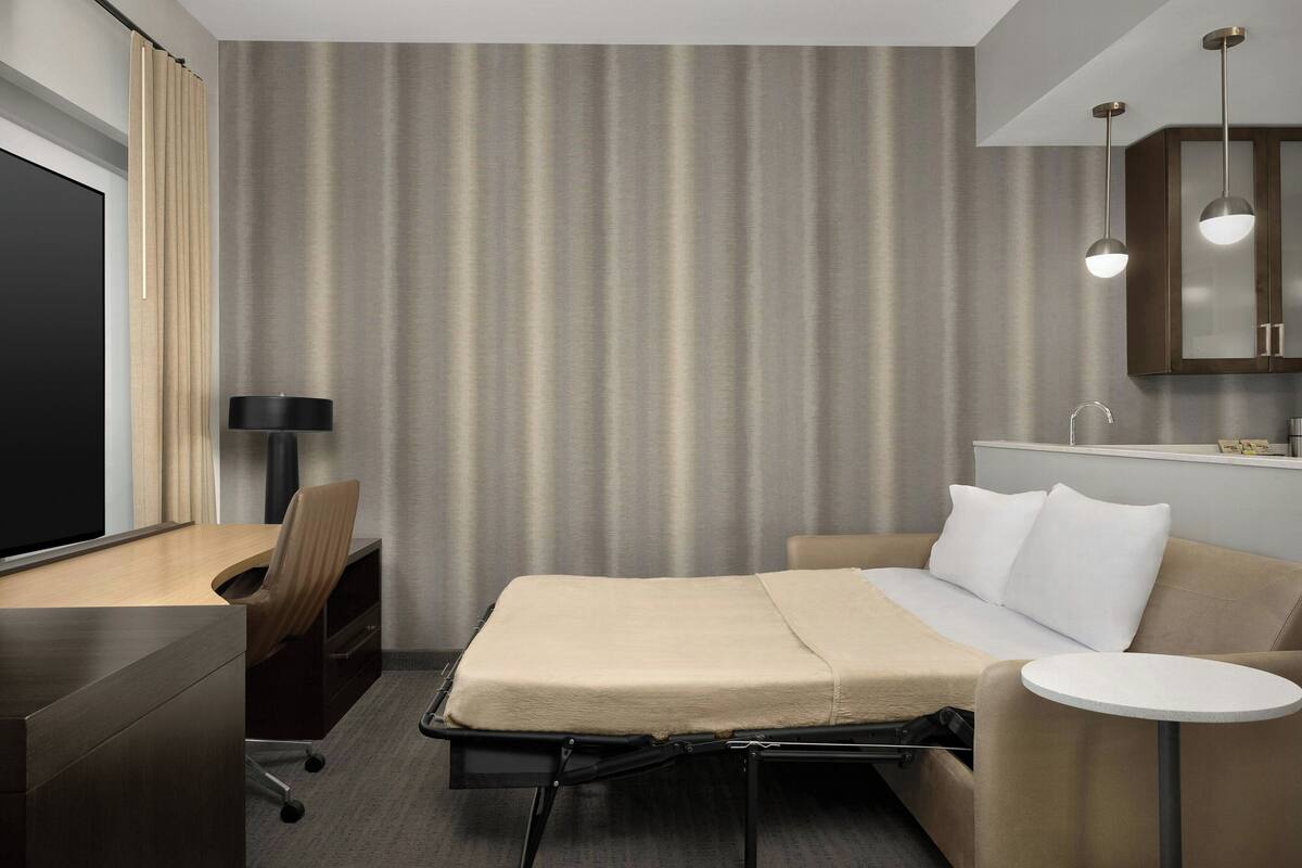 A hotel room with a bed, a desk, a chair, a small table, and a television.