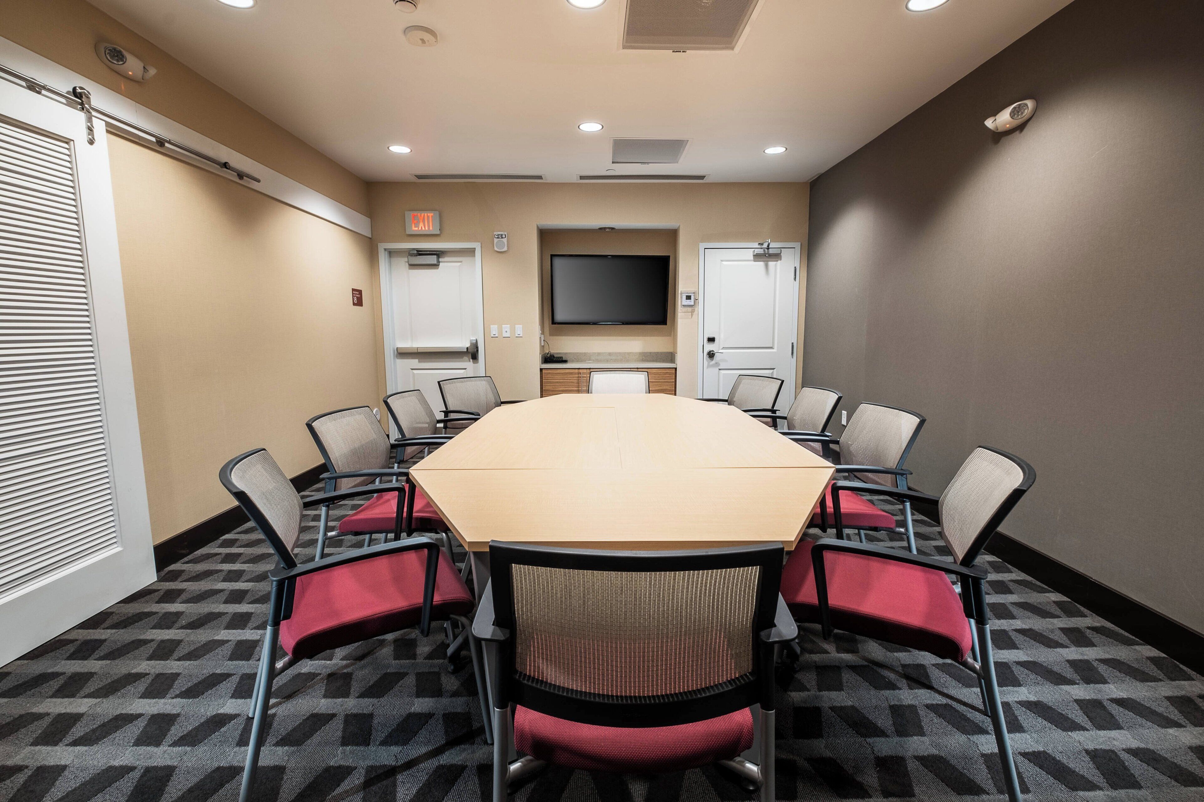 A conference room with a large table, eight chairs, a flat-screen TV, and a door with an exit sign.