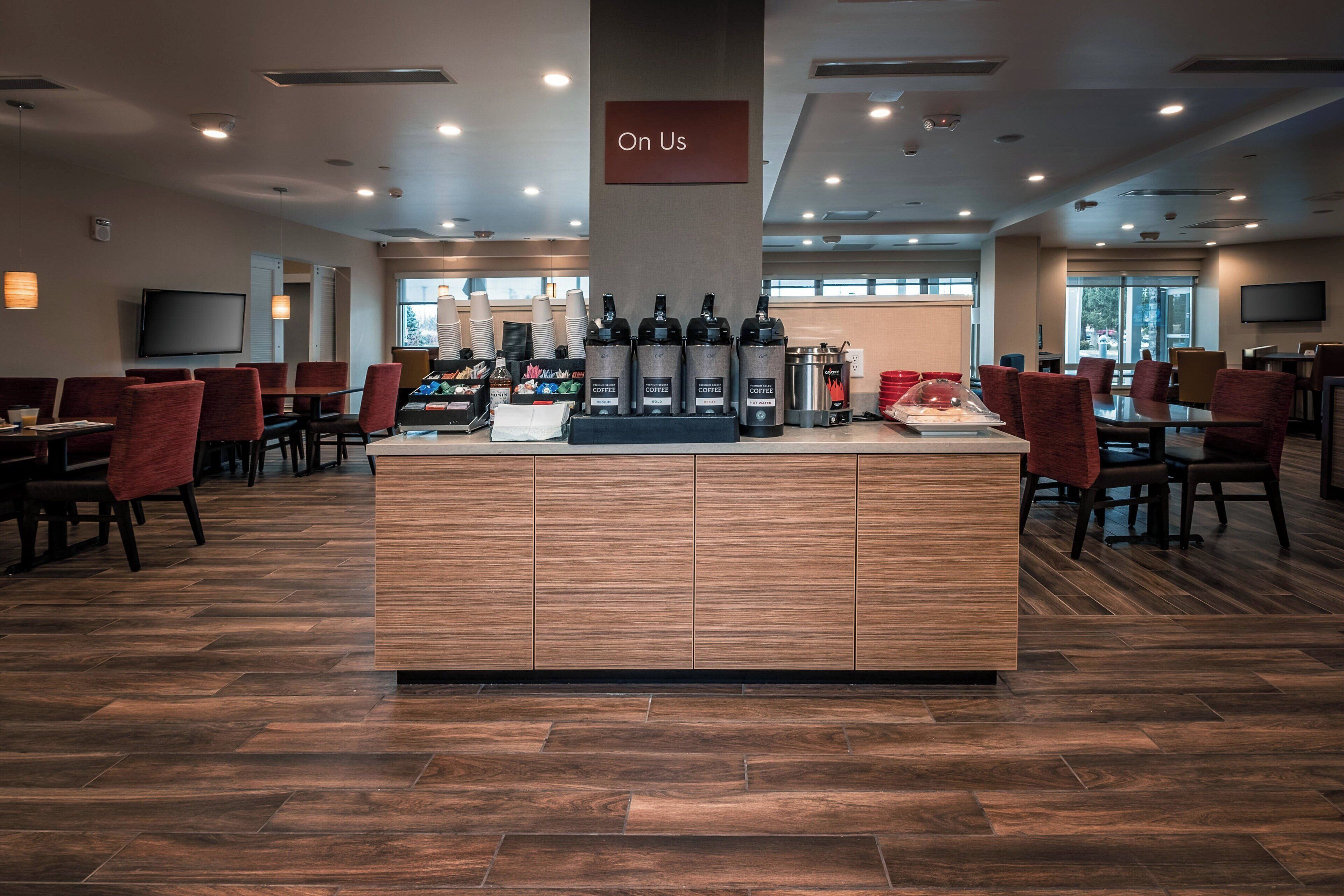 A spacious hotel lobby with a central coffee station, multiple seating areas, and a large television screen on the wall.