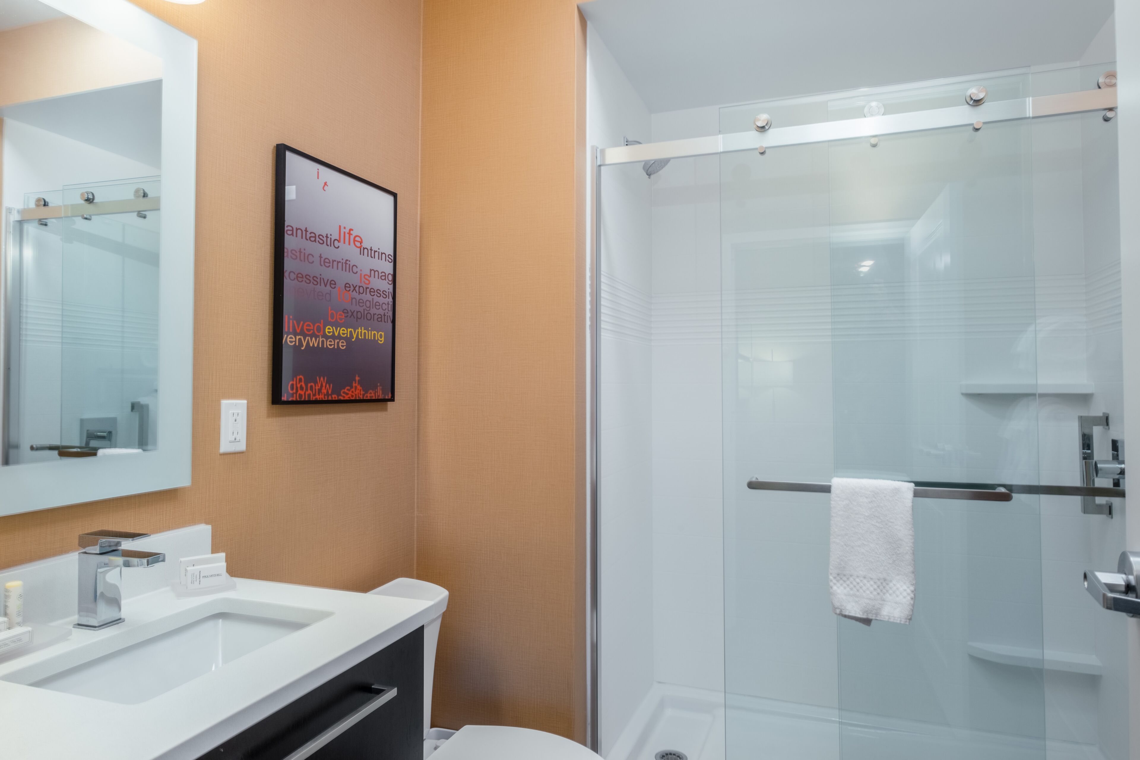 Un baño moderno con un espejo grande, dos lavamanos, una cabina de ducha con mampara de vidrio y un cartel motivacional en la pared.