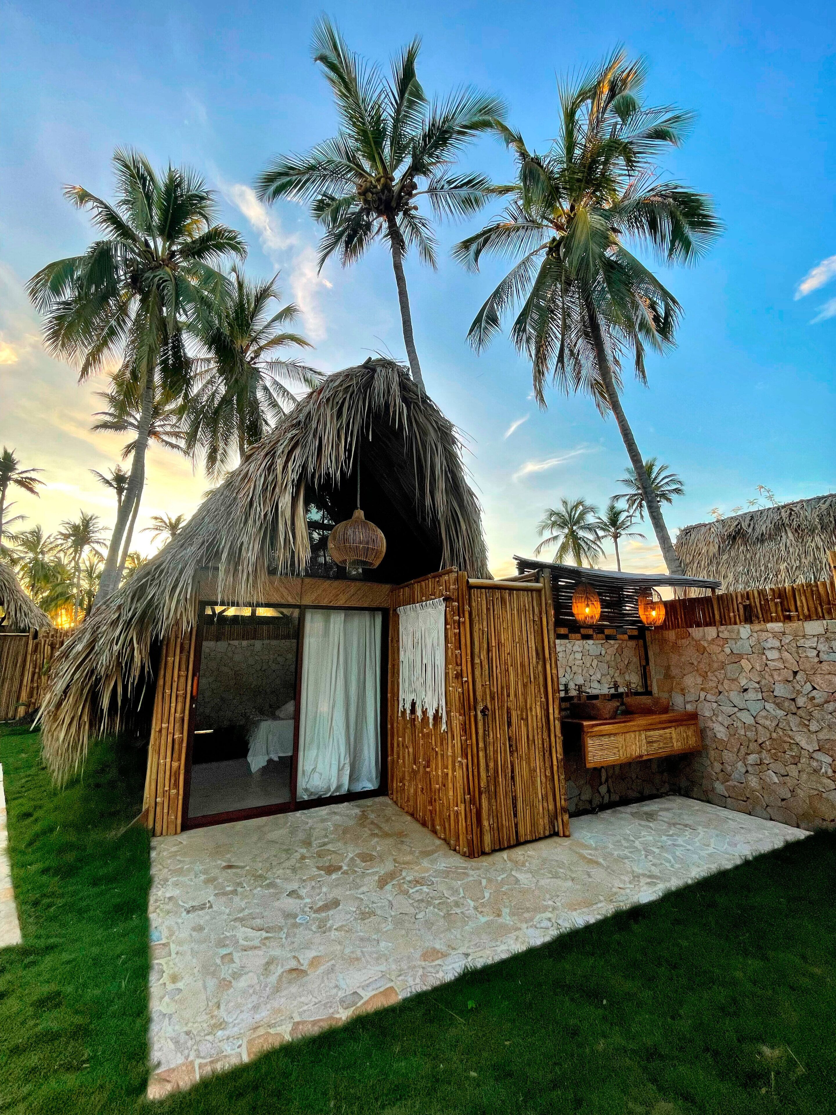 A thatched-roof hut with a stone wall and a wooden bench, surrounded by palm trees.
