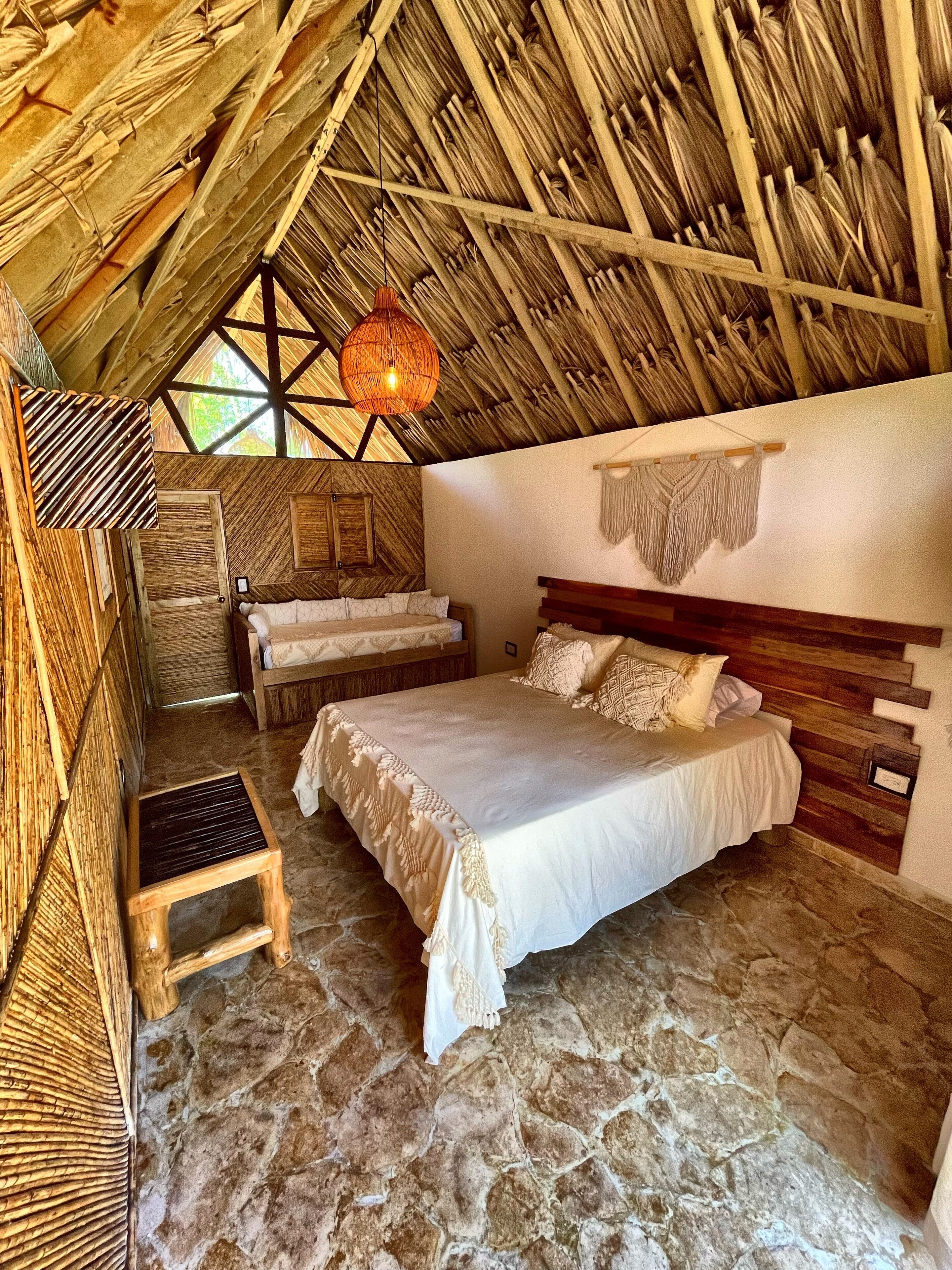 Een rustieke slaapkamer met stenen vloer, een bed met houten hoofdeinde, een klein houten krukje en een geweven plafond.