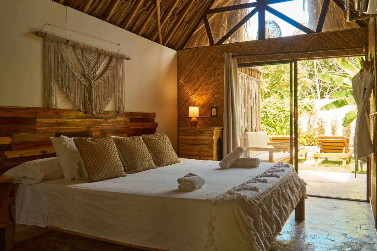 A room with a bed, wooden headboard, macrame wall hanging, and a view of the outdoors.