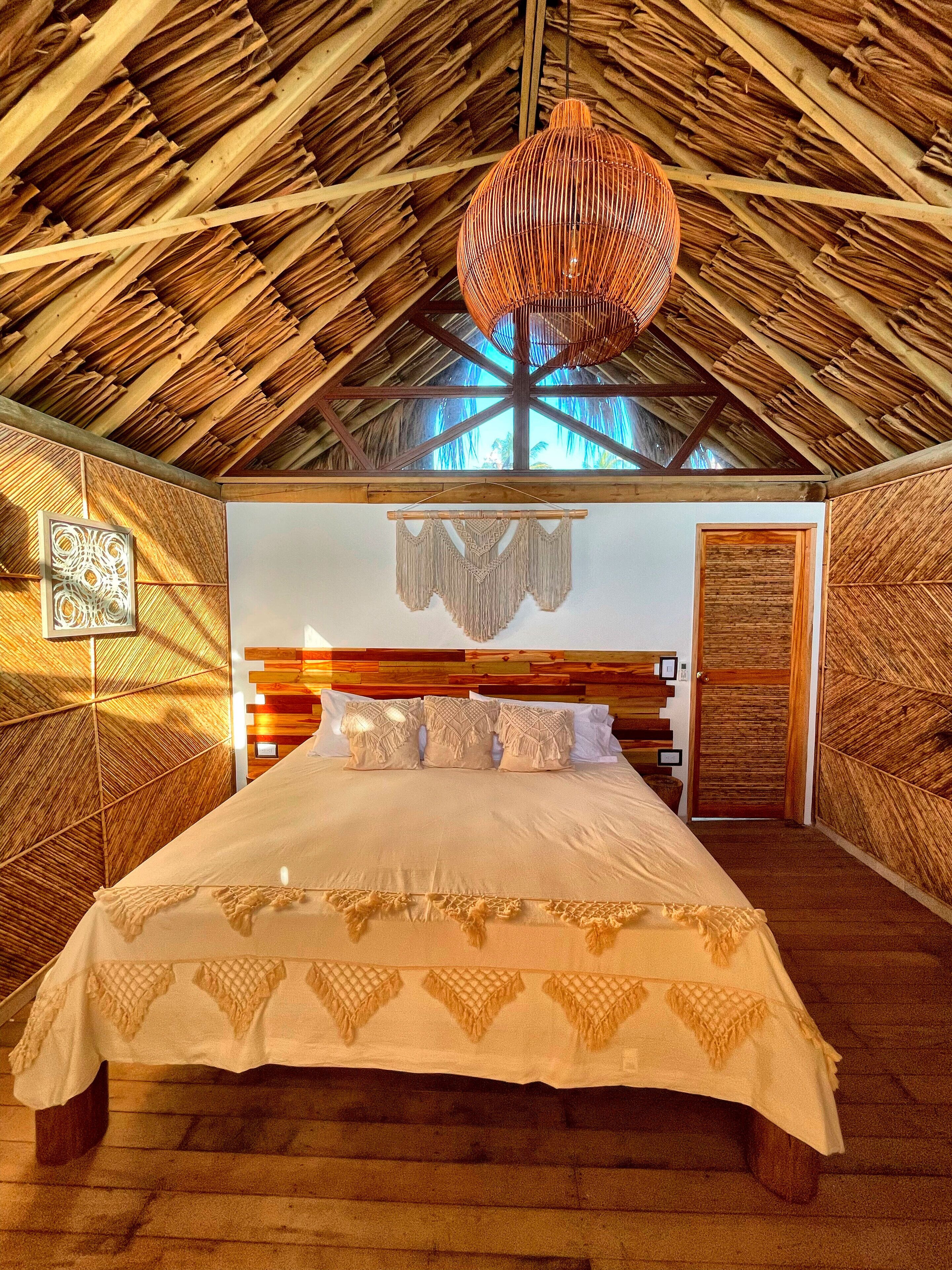 A room with a thatched roof, a bed with a white bedspread, and a macramé wall hanging.