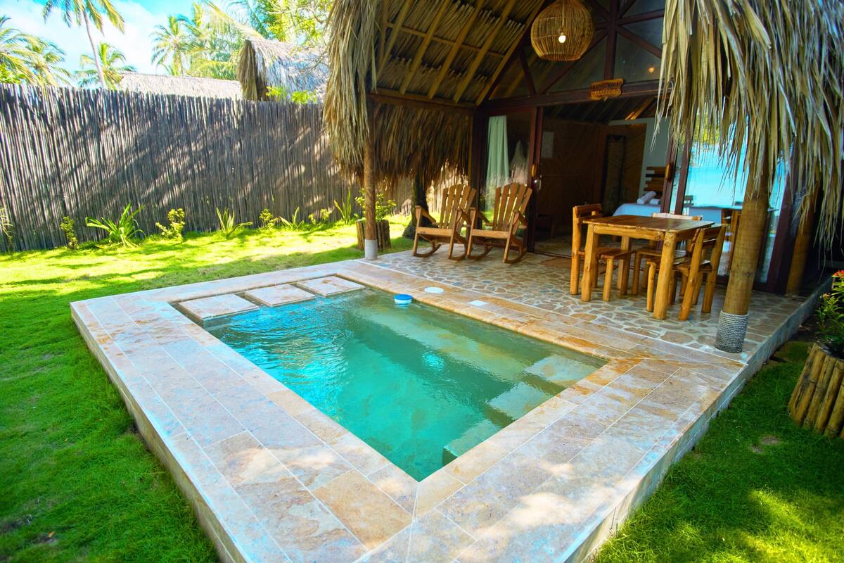 A swimming pool with a stone border, surrounded by a grassy area and a wooden deck with chairs and a table.