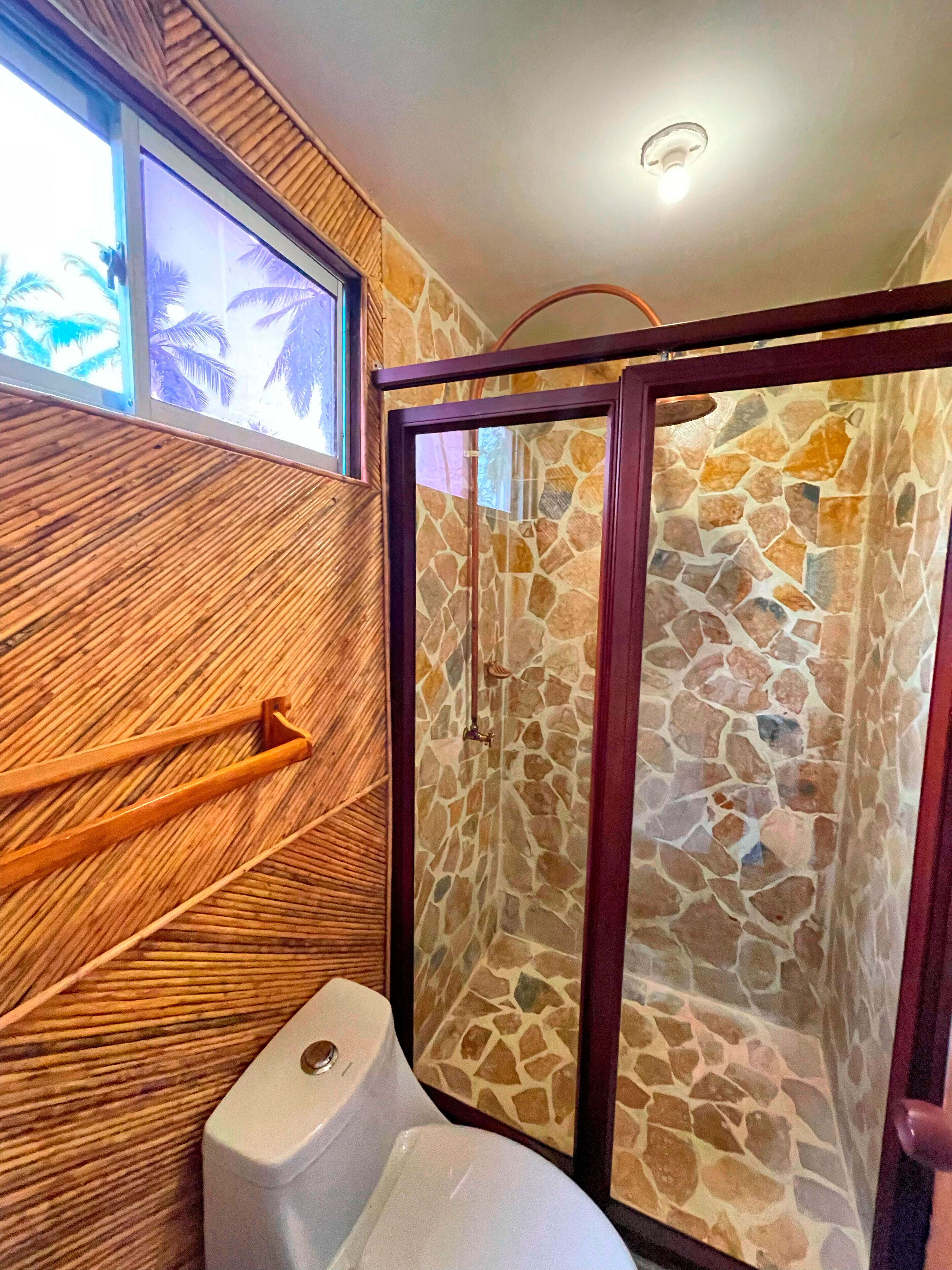 A bathroom with a stone shower enclosure, a wooden wall panel, and a window with a view of palm trees.