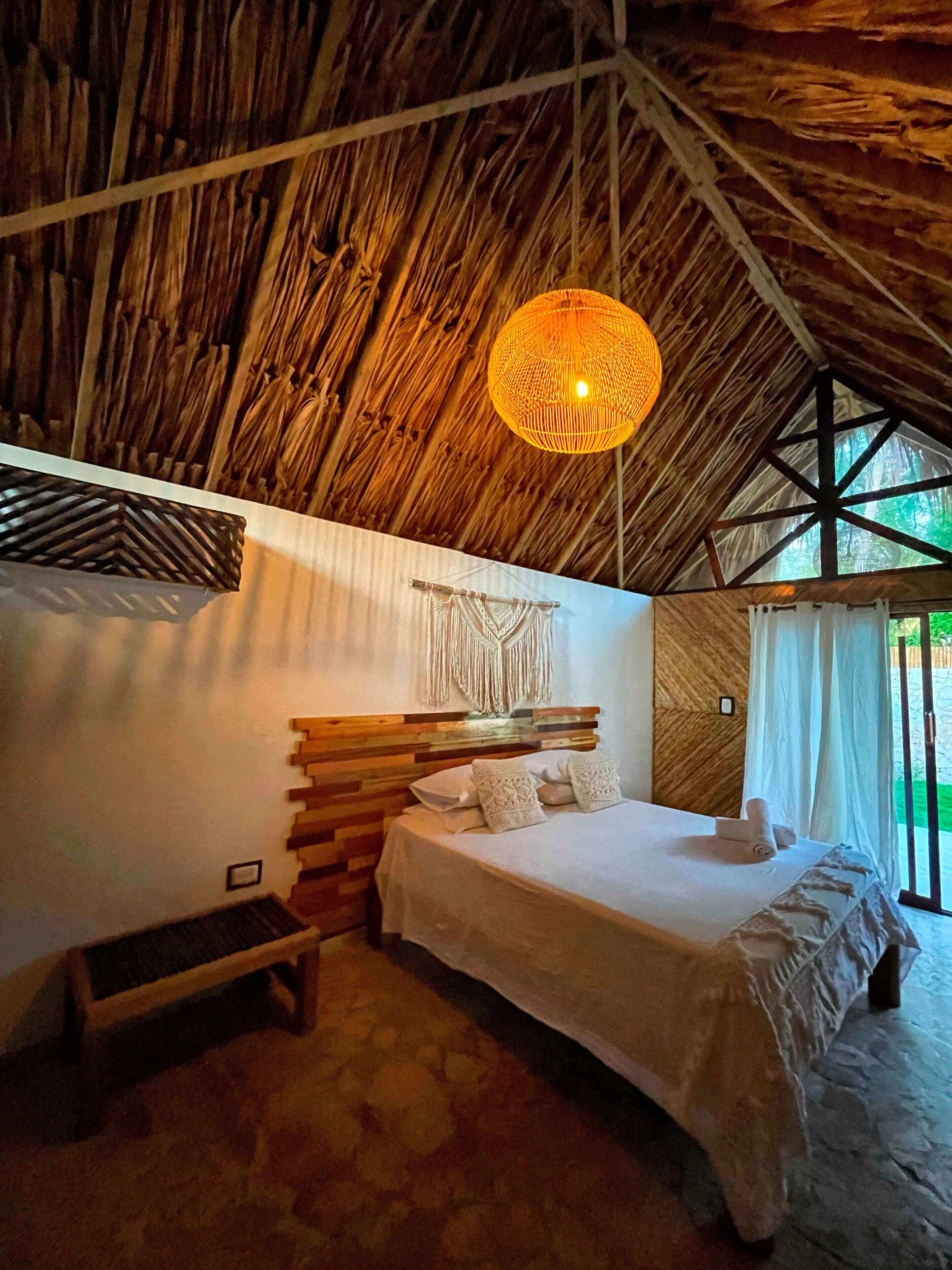 Slaapkamer met een houten schuine plafond, een bed met witte beddengoed en een geweven lamp, met uitzicht op groen buiten.