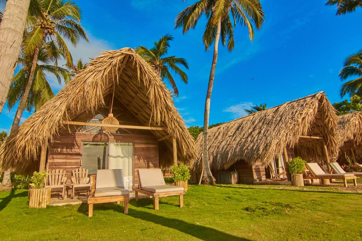 A wooden cabin with a thatched roof, surrounded by palm trees and equipped with lounge chairs and a small table.