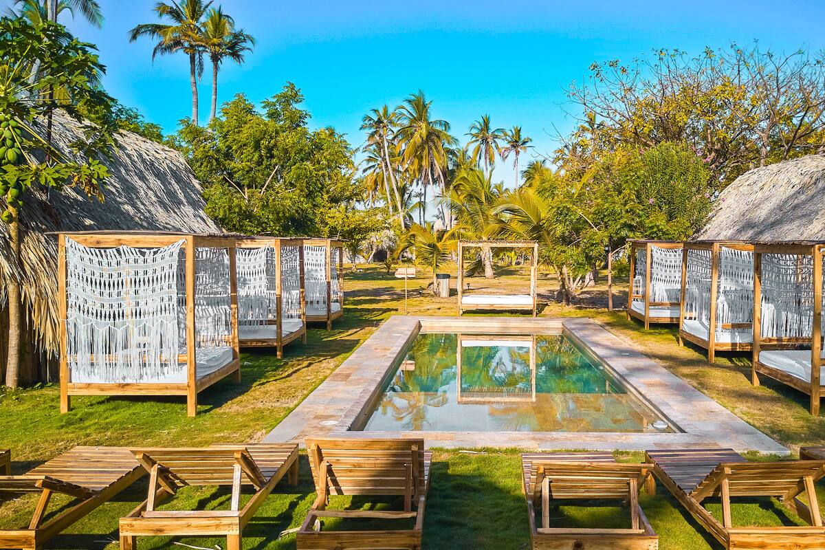 A pool area with wooden loungers and thatched-roof structures, surrounded by palm trees and greenery.