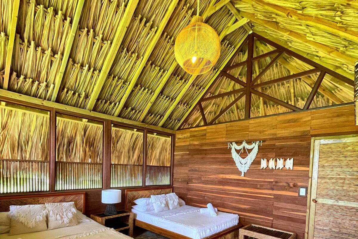 A room with a thatched roof, two wooden beds, a wooden table, and a woven hanging light fixture.