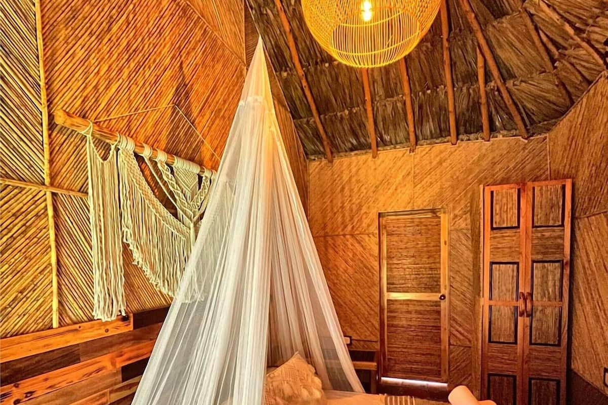 A room with a bed, a hanging macrame chair, a woven light fixture, and wooden doors.