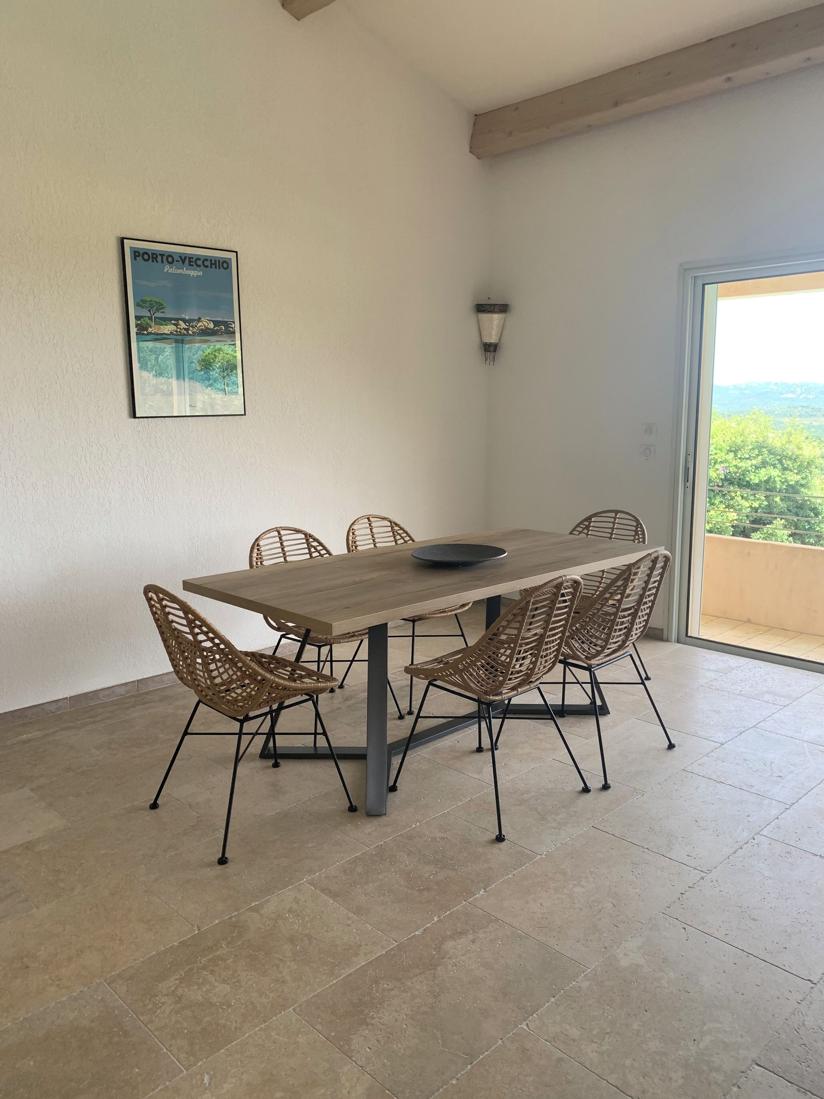 Un comedor con una mesa de madera y sillas de mimbre, una lámpara de pared y un cuadro con una escena costera.