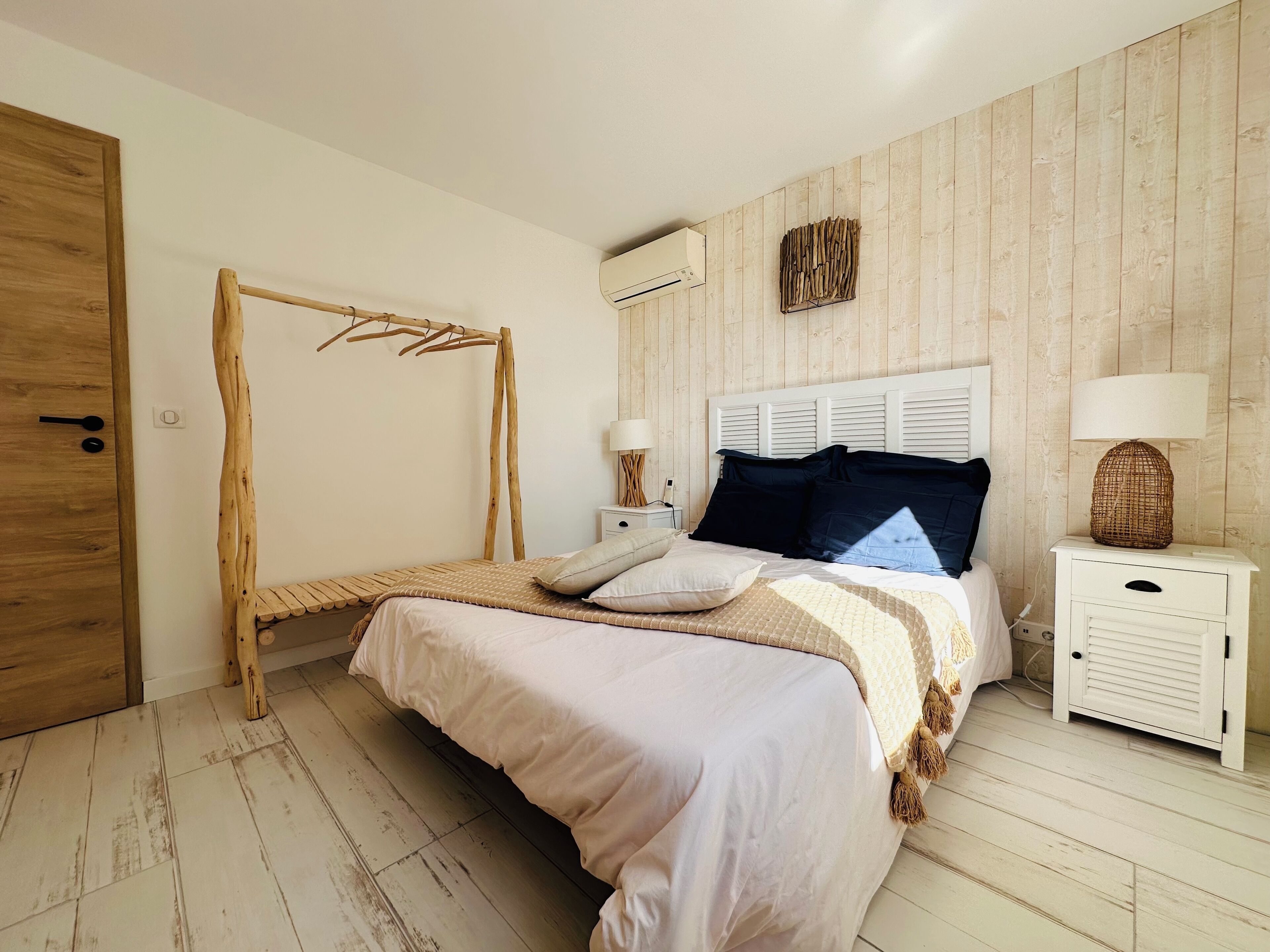 A bedroom with a wooden floor, a bed with a white and beige bedspread, a wooden drying rack, a white bedside table with a lamp, and a wall-mounted air conditioner.