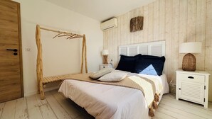 A bedroom with a wooden floor, a bed with a white and beige bedspread, a wooden drying rack, a white bedside table with a lamp, and a wall-mounted air conditioner. - 7 BEDROOM HOUSE WITH AMAZING SEA VIEW AND SWIMMING POOL (Porto-Vecchio)