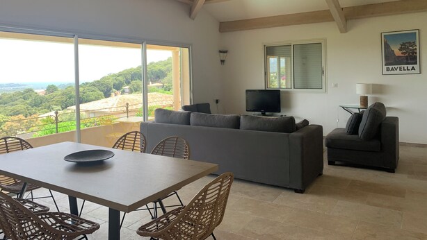 A spacious living area with a large window offering a view of a lush landscape, a grey sofa, a television, and a dining table with wicker chairs. - 7 BEDROOM HOUSE WITH AMAZING SEA VIEW AND SWIMMING POOL (Porto-Vecchio)