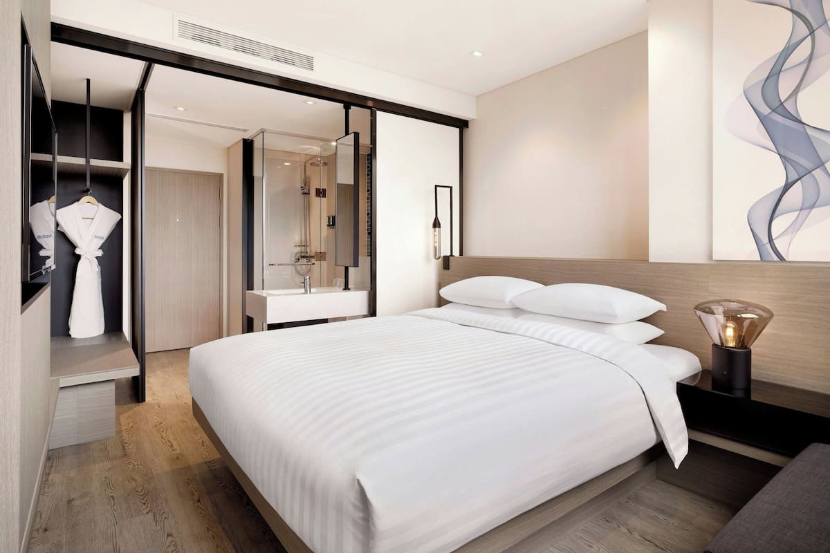 A modern hotel room with a large bed, a walk-in closet, a bathroom with a glass shower, and a wall-mounted light fixture.