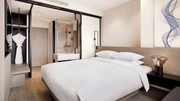 A modern hotel room with a large bed, a walk-in closet, a bathroom with a glass shower, and a wall-mounted light fixture.