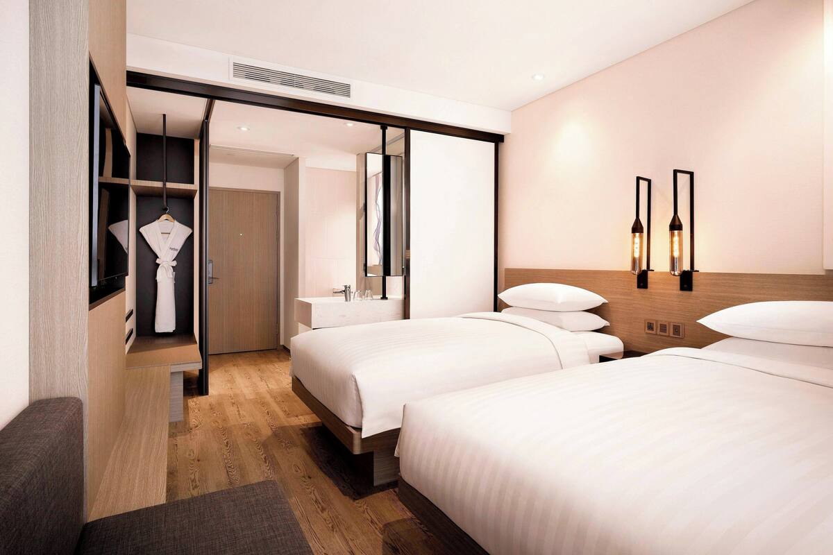 A modern hotel room with two beds, a walk-in closet, a vanity with a mirror, and a wall-mounted light fixture.