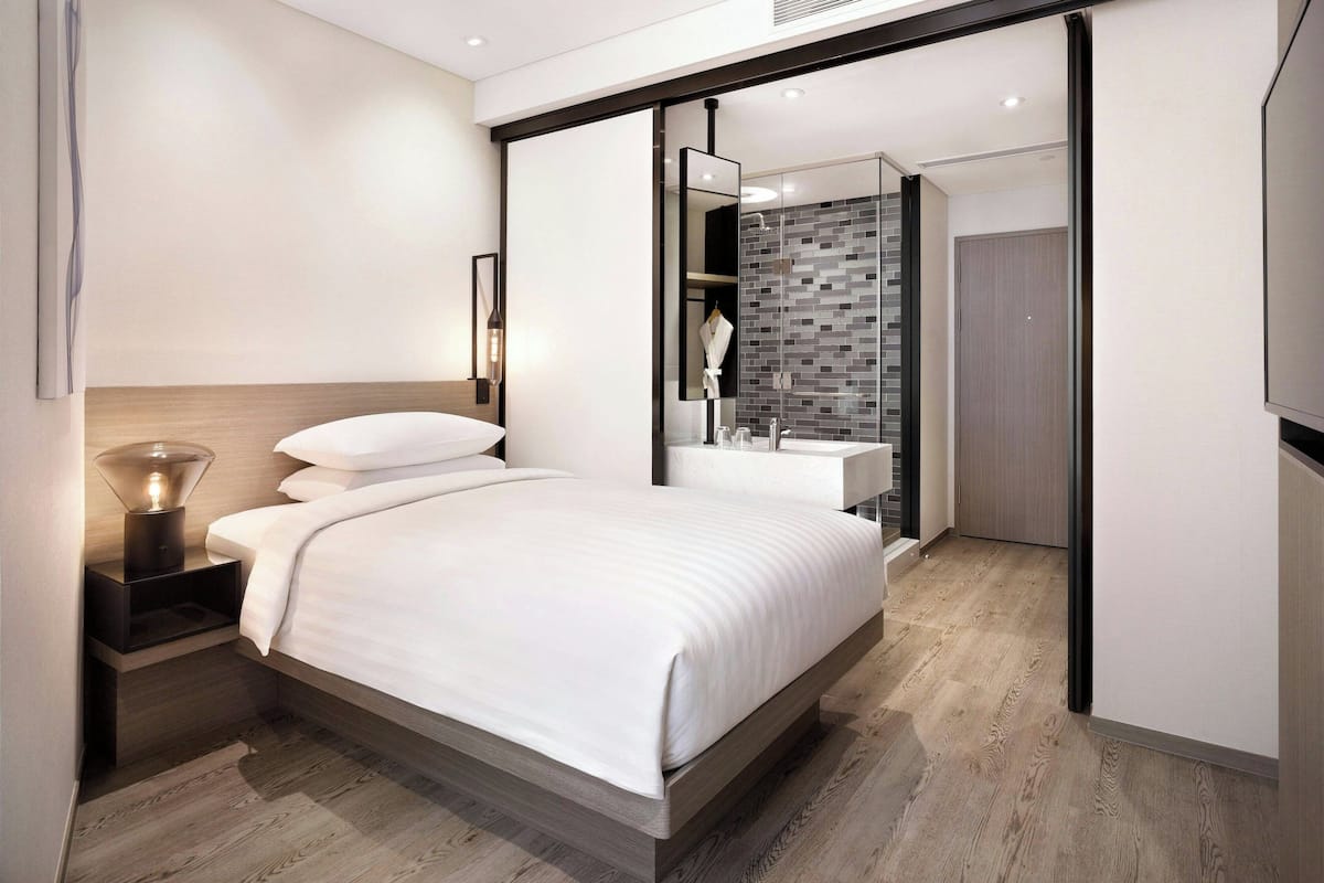 A modern hotel room with a large bed, a bedside table with a lamp, a glass shower enclosure, and a bathroom with a sink and mirror.