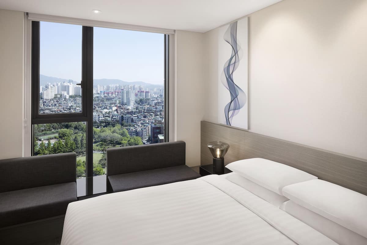 A modern hotel room with a large window offering a city view, a bed with white linens, a gray sofa, and a decorative wall piece.