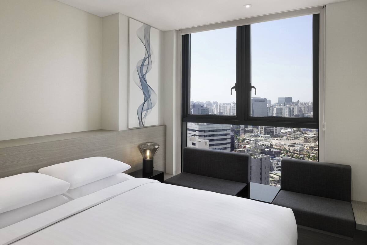 A modern hotel room with a large bed, a bedside table with a lamp, a seating area with a view of the cityscape through a large window.