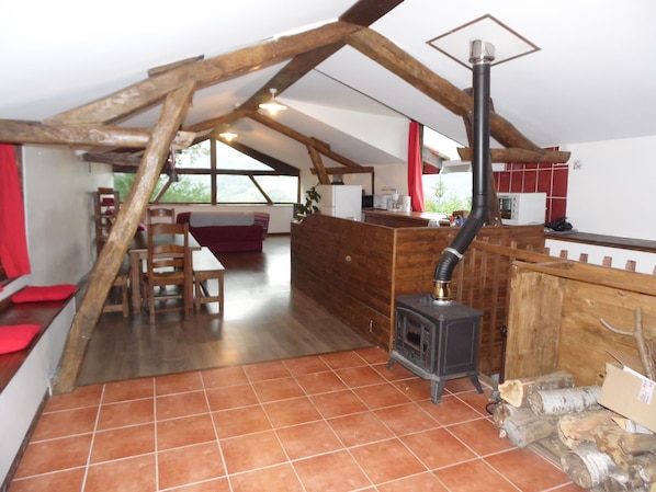 Une cuisine rustique avec un poêle à bois, des poutres apparentes et un coin repas agrémenté d’un canapé rouge.
