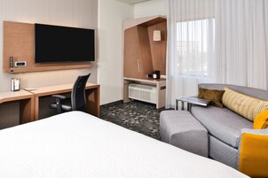 Habitación de hotel con cama, escritorio con silla, televisor, minibar y sofá con cojines.
