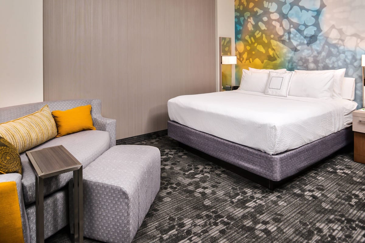 Une chambre d’hôtel avec un lit, un canapé gris agrémenté de coussins jaunes, une petite table et une moquette à motifs.