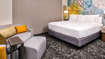 A hotel room with a bed, a gray sofa with yellow cushions, a small table, and a patterned carpet.