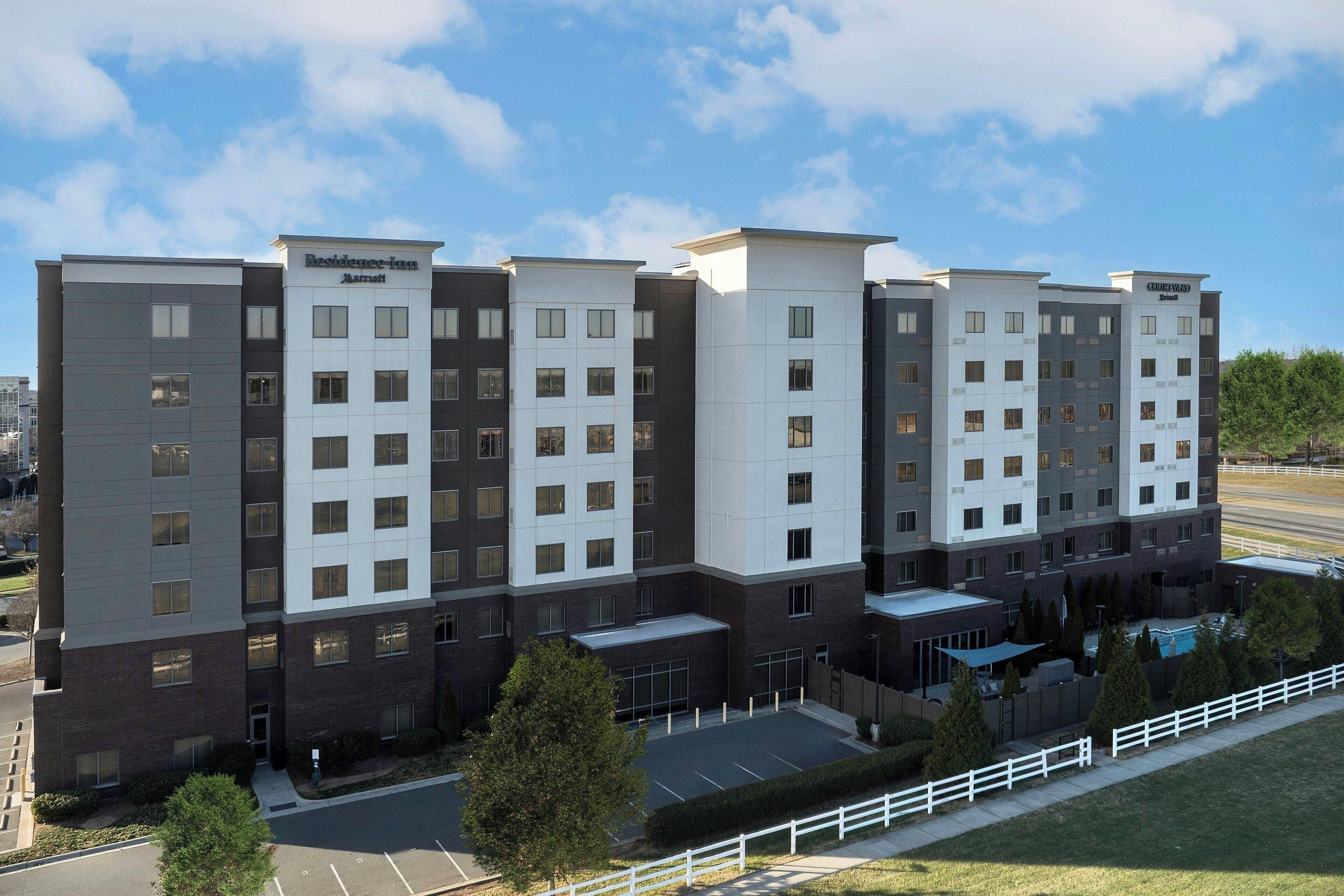 A multi-story hotel with a modern design, featuring a mix of white and dark facade panels, multiple windows per floor, and a parking lot in front.