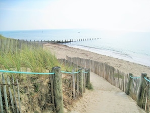 En sandstig som löper längs stranden, med ett trästaket och gräs på vänster sida, och som leder ner till havet.
