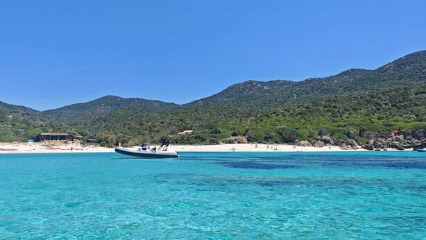 Eine Küstenlandschaft mit klarem türkisblauem Wasser, einem Boot und einem Strand mit Menschen und Gebäuden im Hintergrund.
