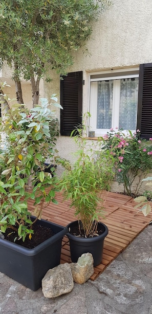 A courtyard with potted plants, a wooden deck, and a building with a window and shutters.
