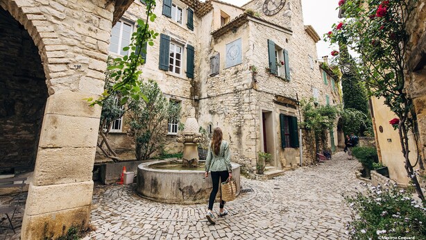 A cobblestone street in a historic town with stone buildings, a fountain, and greenery.