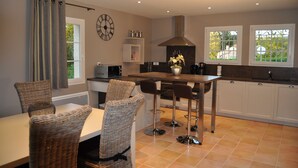 A modern kitchen with a dining area, featuring a central island with bar stools, a clock on the wall, and a view through the windows.