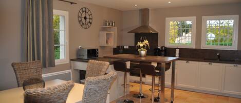 A modern kitchen with a dining area, featuring a central island with bar stools, a clock on the wall, and a view through the windows.